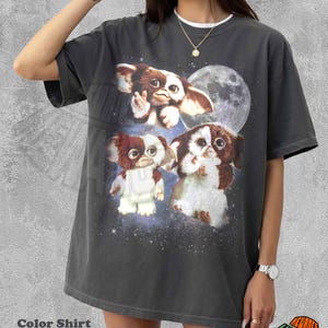 Könnte beinhalten: Ein pfefferfarbenes Kurzarm-T-Shirt mit einem Grafikdruck von drei braun-weißen, pelzigen Kreaturen vor einem dunklen Weltraumhintergrund mit einem großen Mond. Das Design enthält Sterne und einen nebelartigen Effekt um die Figuren.