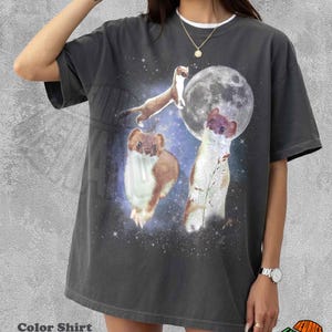 Puede incluir: Camiseta gris oscuro con cuello redondo blanco, que muestra una escena fantástica de comadrejas, luna llena y estrellas. El texto "Color Shirt Pepper" está en la parte inferior. Mangas cortas.