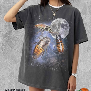 May include: A charcoal gray short-sleeve t-shirt featuring a graphic print of three large insects against a backdrop of a full moon and a starry night sky. The shirt has a crew neckline and a loose, relaxed fit with a faded, washed-out appearance.