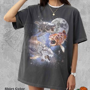 May include: Pepper-colored t-shirt featuring a graphic of three prehistoric fish against a starry background and a full moon. The shirt has a relaxed fit and a white crew neck collar. The text "REWARDS" is visible on the bottom right.