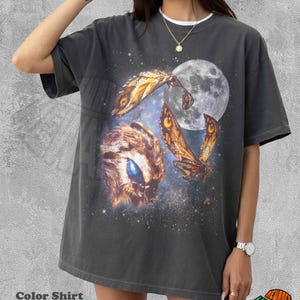 May include: Pepper-colored graphic t-shirt featuring a celestial design with a large moon, moths, and a creature with a blue eye. The shirt has a white collar and the text "Color Shirt Pepper".
