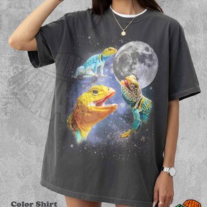 May include: Dark grey t-shirt featuring a vibrant graphic of three lizards against a starry space background with a large moon. The lizards are in shades of yellow, blue, and green. The shirt has the text "Color Shirt Pepper".
