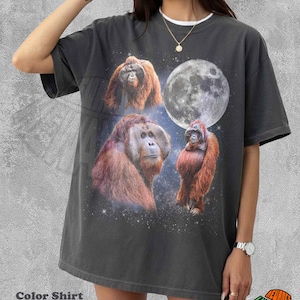 May include: A charcoal gray short-sleeve t-shirt featuring a graphic print of three orangutans set against a starry space background with a large full moon. The shirt has a crew neckline and a relaxed fit, showcasing a detailed cosmic design.