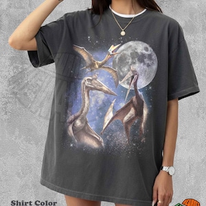 May include: A pepper-colored short-sleeve t-shirt featuring a graphic print of three pterosaurs flying against a backdrop of a large full moon and a starry night sky. The design includes detailed illustrations of the prehistoric flying reptiles.