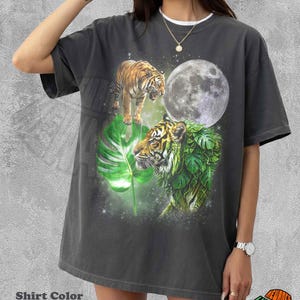 May include: A charcoal grey t-shirt featuring a graphic print of two tigers, one standing and one partially obscured by green monstera leaves, set against a backdrop of a large full moon and a starry night sky. The shirt has a crew neckline.