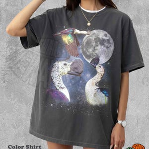 May include: A charcoal gray short-sleeve t-shirt featuring a graphic print of three ducks in a starry night sky with a large full moon. The design includes one duck in flight and two ducks facing each other, set against a dark, cosmic background.