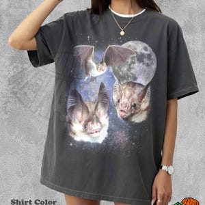 May include: A pepper-coloured short-sleeve t-shirt featuring a graphic print of three bats in flight and resting positions against a backdrop of a large full moon and a starry night sky. The shirt has a crew neckline and a loose, relaxed fit.