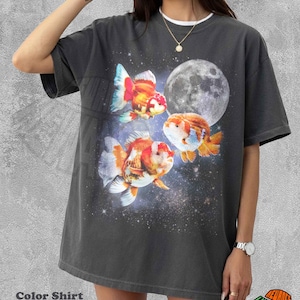 May include: Dark grey t-shirt featuring a celestial design with three colorful goldfish, a full moon, and a starry background. The shirt has a white crew neck and the text "Color Shirt Pepper".