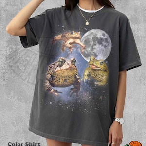 Può includere: T-shirt grigio scuro con una grafica di tre rane su uno sfondo di cielo stellato e luna piena. La maglietta ha il testo "Color Shirt Pepper". La maglietta è di vestibilità ampia.