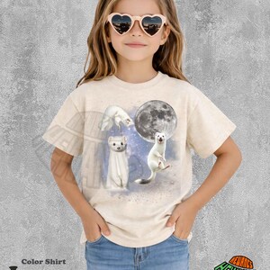 May include: Ivory-colored t-shirt featuring a whimsical design of white ferrets with a full moon. The shirt includes the text "Color Shirt" and "Ivory" at the bottom. The girl is wearing heart-shaped sunglasses and denim shorts.