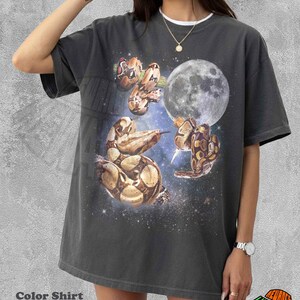 Può includere: Una t-shirt color pepe con una stampa grafica di diversi serpenti avvolti attorno a una luna piena su uno sfondo di galassia stellata blu scuro. La maglietta presenta un girocollo, maniche corte e una vestibilità ampia e rilassata.