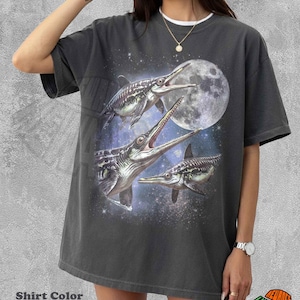 Op de afbeelding: Een peperkleurig t-shirt met een grafische print van drie ichthyosaurussen die zwemmen in een sterrenhemel met een grote volle maan. Het t-shirt heeft een ronde hals en korte mouwen en toont een gedetailleerde illustratie van prehistorische zeereptielen.