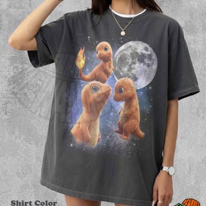 Puede incluir: Una camiseta color pimienta con un estampado gráfico de tres criaturas anaranjadas parecidas a lagartos con ojos grandes. Una criatura tiene una llama en la cola. El fondo muestra una luna llena y un cielo nocturno estrellado.
