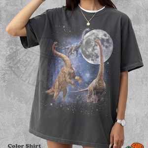 Pode incluir: Uma t-shirt de manga curta na cor pimenta com uma estampa gráfica de três dinossauros de pescoço comprido numa cena espacial estrelada com uma lua cheia grande. O design inclui um fundo cósmico com estrelas dispersas e nuvens.