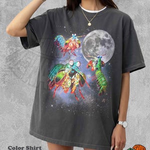 May include: A charcoal gray short-sleeve t-shirt featuring a graphic print of three colorful mantis shrimp floating in a starry space scene with a large moon. The shirt has a crew neckline and a relaxed fit, showcasing vibrant red, green, and blue hues.