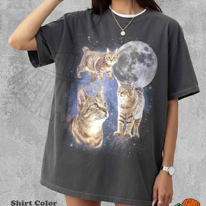 May include: A pepper-colored short-sleeve t-shirt featuring a graphic print of three brown tabby cats against a dark blue space background with a large full moon. The design includes a starry sky motif surrounding the cats and the moon.