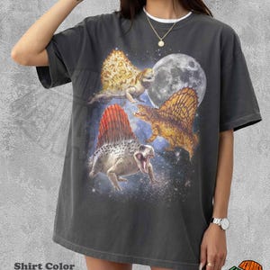 May include: A pepper-coloured short-sleeve t-shirt featuring a graphic print of three dimetrodon dinosaurs in a space setting with a large moon. The design includes a starry nebula background and the dinosaurs are depicted in shades of brown and orange.