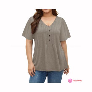 May include: A taupe-colored top with a V-neckline and short, flared sleeves. The shirt features a series of vertical pleats down the front, secured by three dark buttons. The bottom of the shirt flares out slightly. The model is wearing blue jeans.
