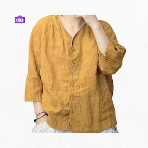 Può includere: Camicia di lino giallo senape con scollo a V e chiusura con bottoni. La camicia ha maniche a tre quarti e una vestibilità ampia. L'immagine include anche una piccola grafica viola e bianca "SPEDIZIONE GRATUITA" nell'angolo in alto a sinistra.