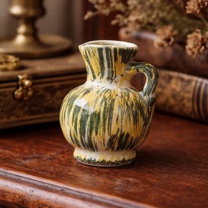 May include: A small, decorative ceramic pitcher with a handle. The pitcher is painted with vertical strokes of yellow and green over a white base. The pitcher sits on a dark wooden surface.