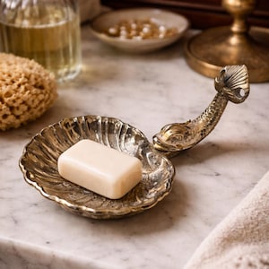 以下が含まれることがあります： 貝殻の形をした真鍮製の装飾的な石鹸置き。石鹸が置かれています。石鹸置きは魚の形をしたハンドルが付いています。大理石の表面に置かれています。背景にはスポンジとボトルがあります。