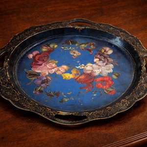 May include: A vintage, circular serving tray with a blue central area and a floral pattern. The tray has a scalloped edge and two handles. The floral design features red, pink, purple, and yellow flowers. The rim is black with gold detailing.