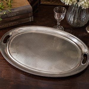 Pode incluir: Uma bandeja de servir oval de metal prateado com um padrão texturizado de círculos concêntricos na base, uma borda estilo corda e duas alças integradas nas extremidades opostas. A bandeja tem um acabamento polido e uma borda elevada.