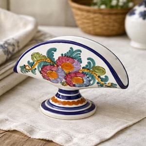 May include: A ceramic napkin holder with a white base, adorned with a blue rim and a floral design in shades of pink, purple, and orange. The holder has a curved shape and sits on a pedestal base with blue and orange stripes. A decorative kitchen item.