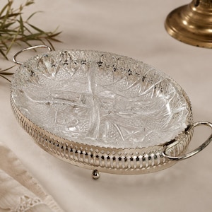 Op de afbeelding: Een ovale, verdeelde serveerschaal met een helder, geslepen glazen interieur en een zilverkleurige metalen rand en handvatten. De schaal heeft een decoratief, gestructureerd patroon en wordt verhoogd door kleine voetjes.