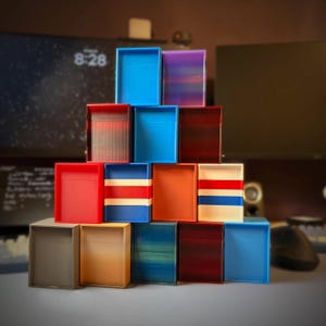 May include: A pyramid stack of fourteen rectangular open-top storage boxes in various solid colors and striped patterns, including shades of blue, red, orange, beige, gray, and purple, arranged in a four-level structure on a flat surface.