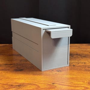 May include: A light gray rectangular storage box with a smooth finish and a horizontal handle on the front panel. The box features a clean, boxy design with a flat top surface and a recessed front drawer section, constructed with visible layered lines.