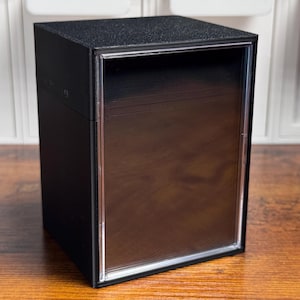 May include: A rectangular black storage box with a textured matte finish and a clear transparent front panel. The container features a vertical orientation with a solid black top and sides, designed to hold items visible through the front window.