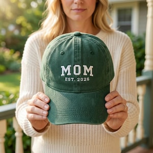 Könnte beinhalten: Dunkelgrüne Baseballkappe mit dem Wort "MOM" in weißen Blockbuchstaben gestickt. Unter dem Wort "MOM" steht der Text "EST. 2026". Die Kappe ist aus einem weichen Material gefertigt und hat einen gebogenen Schirm.