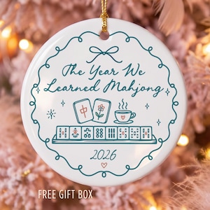 May include: A round white ceramic ornament featuring a teal scalloped border and the text The Year We Learned Mahjong. The design includes illustrations of two mahjong tiles, a teacup, a rack of tiles, and the year 2026 with a small heart.