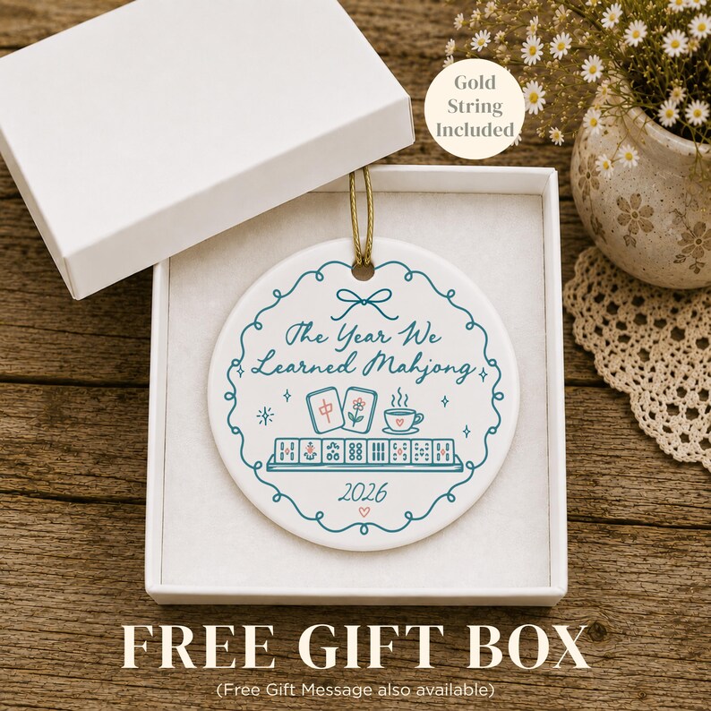May include: A round white ceramic ornament with a gold string, presented in a white gift box. The ornament features a teal scalloped border, the text "The Year We Learned Mahjong", a mahjong tile illustration, a teacup, and the year 2026.