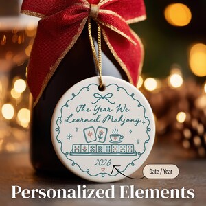 May include: A round white ceramic ornament hanging by a gold string from a bottle neck with a red ribbon. The ornament features a teal decorative border, the text "The Year We Learned Mahjong", illustrations of mahjong tiles and a cup, and "2026".