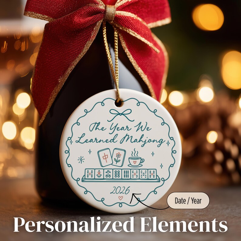 May include: A round white ceramic ornament hanging by a gold string from a bottle neck with a red ribbon. The ornament features a teal decorative border, the text "The Year We Learned Mahjong", illustrations of mahjong tiles and a cup, and "2026".