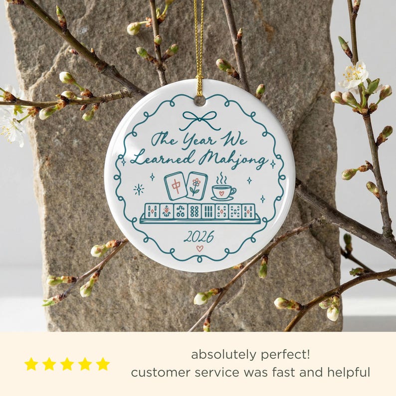 May include: A round white ceramic ornament with a gold string, featuring a teal decorative border and the text "The Year We Learned Mahjong" above illustrations of mahjong tiles, a teacup, and the year "2026" with a small red heart.