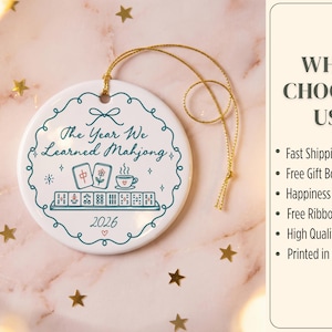 May include: A round white ceramic ornament with a gold hanging string. The design features a teal scalloped border, the text "The Year We Learned Mahjong", a graphic of mahjong tiles and a teacup, and the year "2026" with a small red heart.