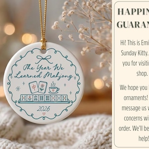 May include: A round white ceramic ornament hanging from a gold string. The front features a teal scalloped border, the text "The Year We Learned Mahjong", illustrations of mahjong tiles and a teacup, and the year "2026" with a small heart.