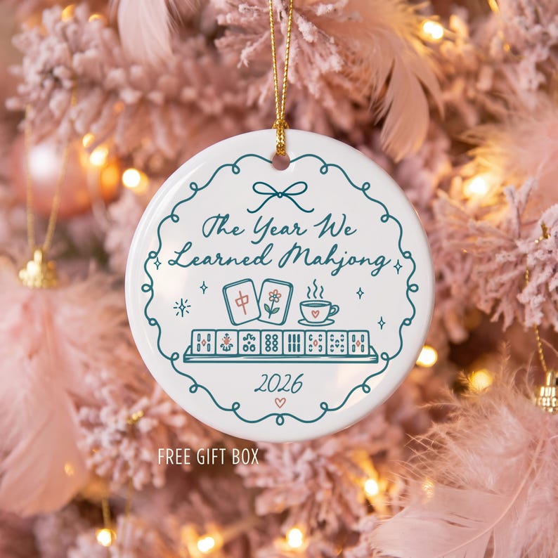 May include: A round white ceramic ornament featuring a teal scalloped border and the text The Year We Learned Mahjong. The design includes illustrations of two mahjong tiles, a teacup, a rack of tiles, and the year 2026 with a small heart.