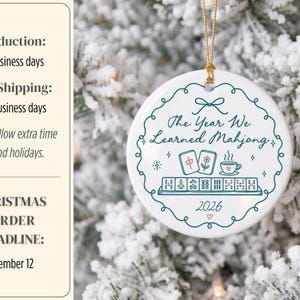 May include: A round white ceramic ornament hanging by a gold string. The design features a teal scalloped border, the text The Year We Learned Mahjong, illustrations of mahjong tiles and a teacup, and the year 2026 in teal and red ink.