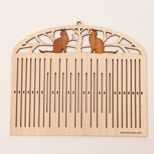 May include: A wooden weaving loom with a decorative top featuring two brown cat silhouettes perched on a tree branch design. The loom has vertical slots and a small hole at the top for hanging. The website address www.jumaka.com is visible at the bottom.