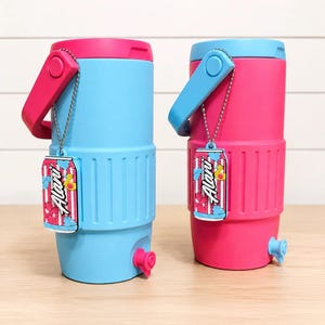 May include: Two insulated water coolers, one blue and one pink, with matching handles and lids. Each cooler has a spigot and a decorative tag with the text "Alani". The coolers are designed to keep beverages cold.