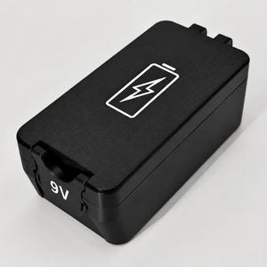 May include: A rectangular black 9V battery with a matte finish. The top surface features a white graphic icon of a battery with a lightning bolt inside. The front edge displays the white text 9V clearly printed on the casing of the battery.