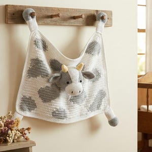 Può includere: Una coperta per bambini all'uncinetto fatta a mano con un design a mucca. La coperta bianca ha macchie grigie e una testa di mucca 3D al centro. La coperta è appesa a un attaccapanni di legno con pioli. Un oggetto morbido e accogliente.
