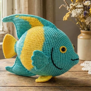 May include: A handmade, crocheted fish in shades of teal and yellow. The fish has a rounded body with fins and a tail. The fish is sitting on a wooden surface. The fish has a smiling face with black details.