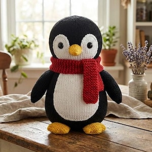 May include: A knitted penguin toy with a black body, white belly, and a red scarf. The penguin has yellow feet and a yellow beak. The toy is on a wooden table.