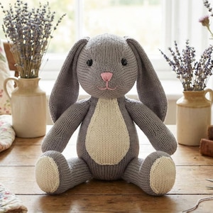 Peut inclure: Un jouet lapin en tricot gris avec de longues oreilles, un nez rose et un ventre blanc. Le lapin est assis sur une surface en bois. L'arrière-plan comprend des vases avec des fleurs et une courtepointe à motifs.