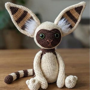 May include: A handmade crochet plush toy in the shape of a lemur. The toy features large, striped ears, a brown face, and a long, striped tail. The body is cream-colored with brown accents and green eyes. The toy is sitting on a wooden surface.